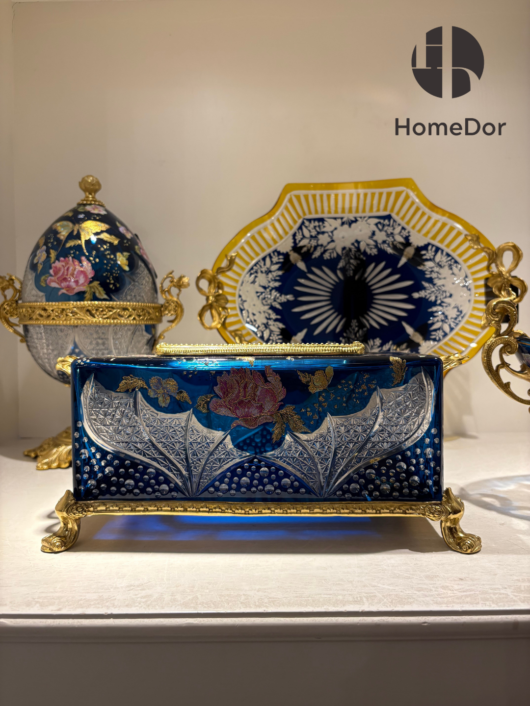 Inspired by the romance of Belle Époque decorative arts, each piece combines luminous Aurora Glass with intricate gilt mounts, creating a harmonious balance between elegance and imagination. The rich blue palette evokes twilight skies, while the golden floral motifs bring warmth and vitality, as though illuminated by a hidden moonlit glow.
Displayed together, the collection creates the atmosphere of a secret garden preserved in glass—one where flowers never fade, butterflies never depart, and beauty linger