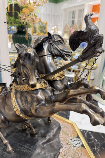 Two horses rear mid-stride, reins pulled taut as a helmeted charioteer leans into the motion behind them. The composition captures a single frozen instant — hooves still airborne, the chariot wheel caught turning — rendered in deep bronze patina with gilt picked out along the harness, the wheel spokes, and the warrior's armor.