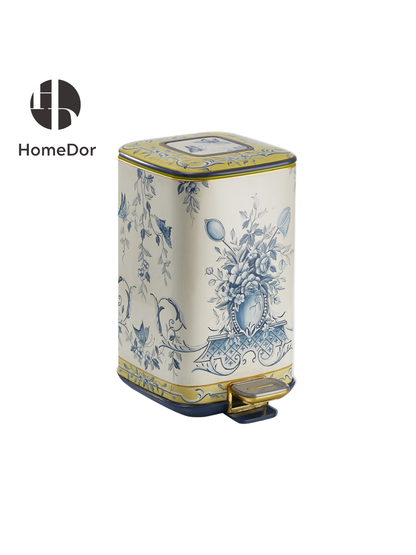 A cream pedal bin painted in soft blue florals, a garland of leaves and blossoms wrapped around a central urn motif. The linework is fine and continuous, closer to a toile print than a bold pattern, with a thin gold trim running along the top and base.