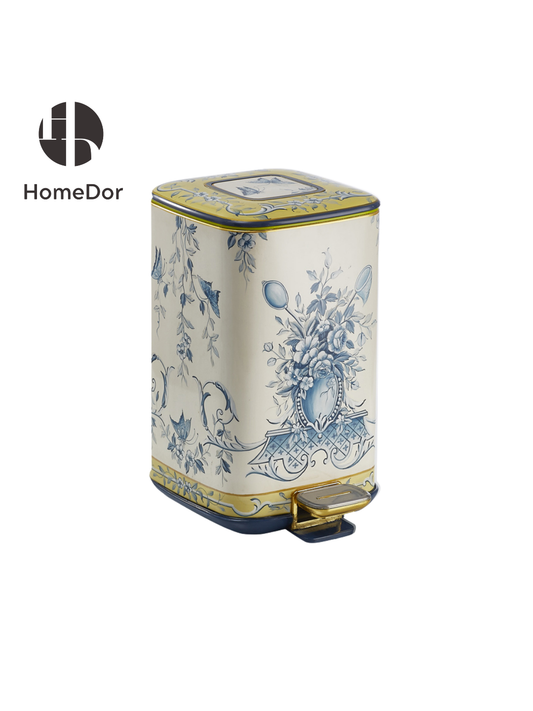 A cream pedal bin painted in soft blue florals, a garland of leaves and blossoms wrapped around a central urn motif. The linework is fine and continuous, closer to a toile print than a bold pattern, with a thin gold trim running along the top and base.