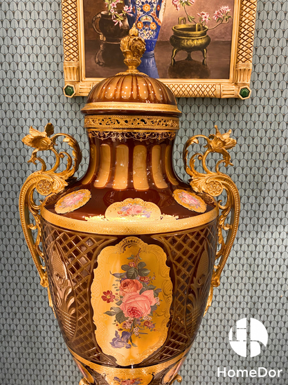 Crowned by a richly ornamented domed lid and framed by elaborate ormolu-style handles, the urn is adorned with hand-painted floral medallions set against a luminous amber-toned ground. Delicate latticework, gilded detailing, and jewel-like decoration enrich every surface, creating layers of visual depth that reveal new details from every angle.